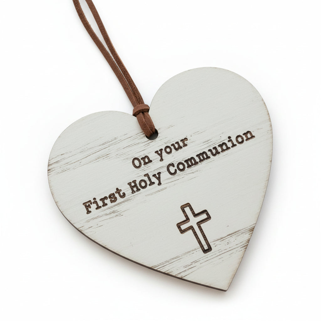 A wooden heart-shaped decoration with a cross and the text 'On your First Holy Communion' inscribed on it. It is hanging from a brown cord.