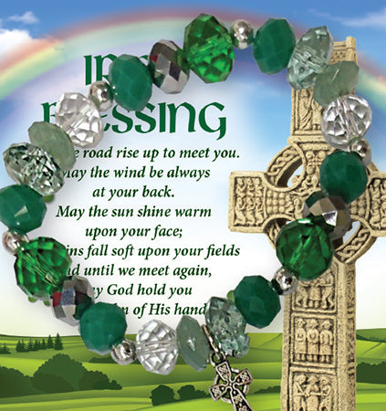 A glass bracelet with green and clear beads, featuring a metal motif in the shape of a cross, displayed in front of a card with an Irish blessing.