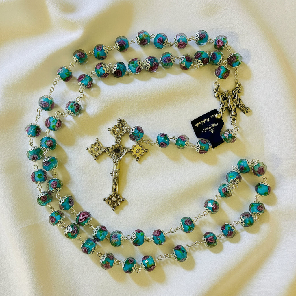 A rosary with turquoise beads and pink rose accents, featuring a silver cross and detailing, laid out on a white surface.
