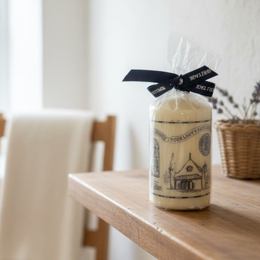A small white candle with a black illustrated label, wrapped in clear cellophane and tied with a black ribbon, sitting on a brown countertop.