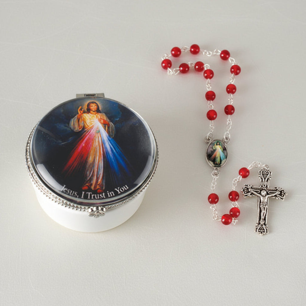 A porcelain box with a picture of Jesus in a glass dome on top, alongside a ruby acrylic rosary with a cross and red beads.