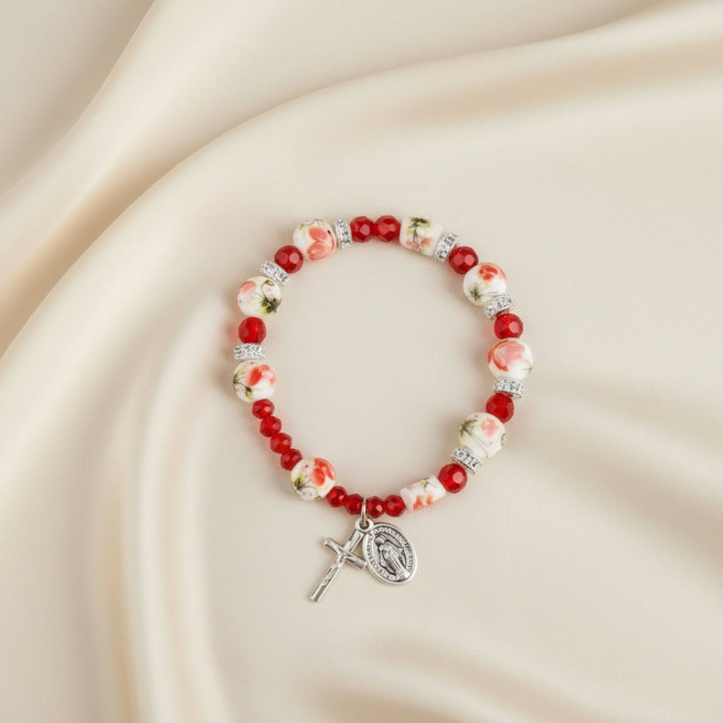A bracelet with red and white beads, glass and ceramic components, and a floral design on the ceramic beads. It includes a metal crucifix and miraculous medal, packaged under the brand 'Inspirations'.